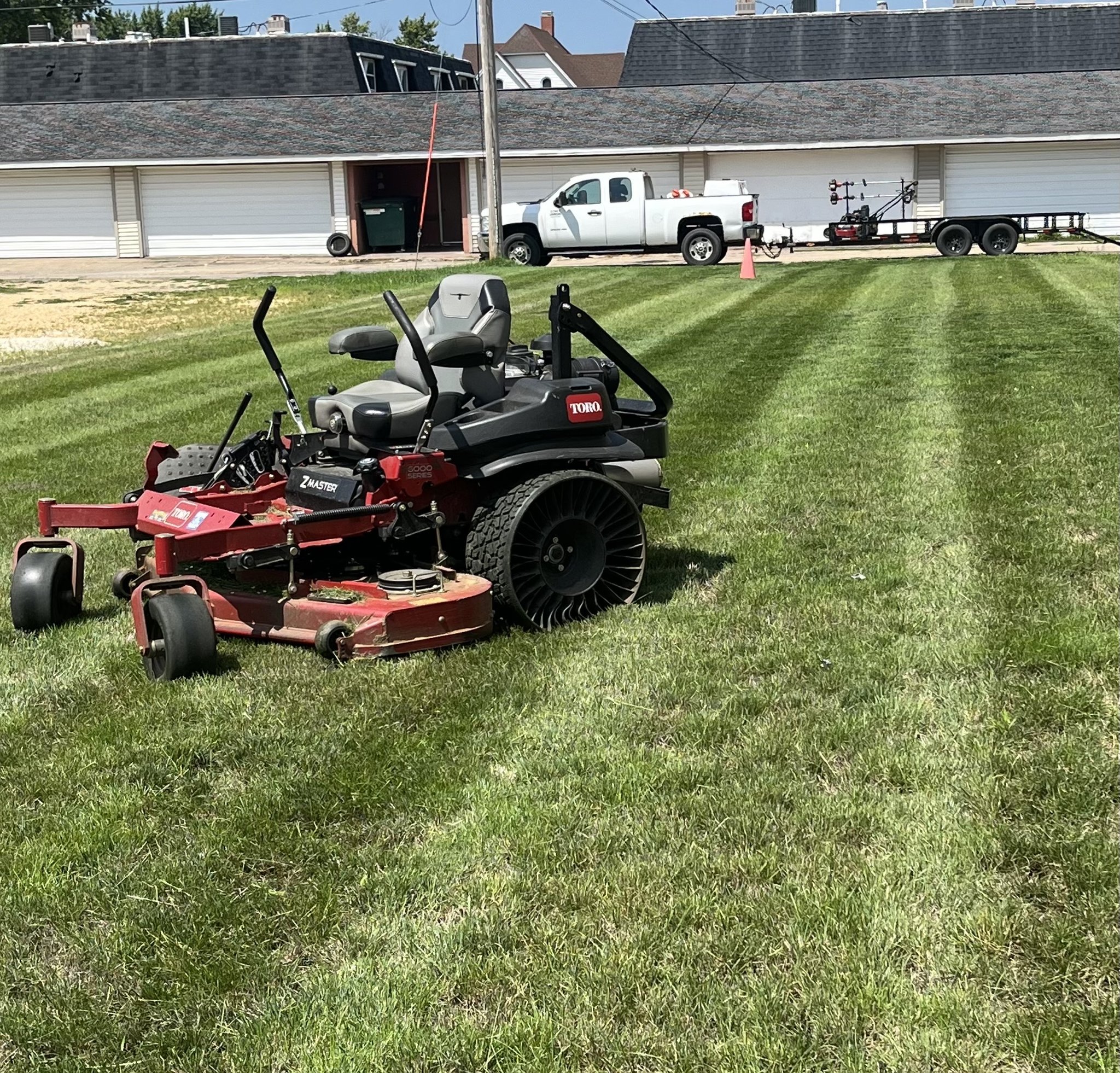 Toro zero-turn mower cutting crisp stripes into a commercial lawn with company truck and trailer in the background